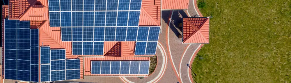 Aerial view of a building with extensive blue solar panels on a red-tiled roof and curved pathways beside a green lawn.