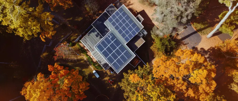 Aerial view of a modern house with solar panels on the roof, surrounded by vibrant autumn trees in orange and yellow.