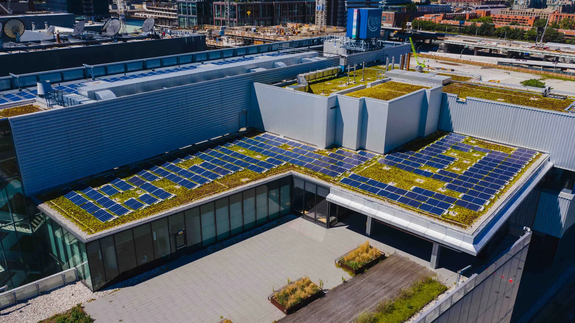 Rooftop solar panels cover a modern building terrace with green roof patches and urban surroundings in the distance.
