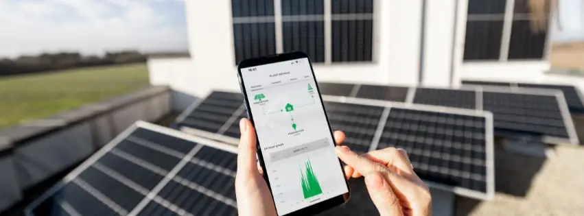 Person holding a smartphone displaying a solar monitoring app, with multiple rooftop solar panels in the background.