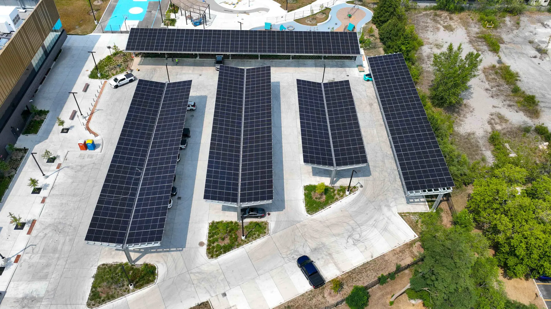 Aerial view of multiple long solar panel carport arrays arranged around a concrete parking area, with a few vehicles and surrounding greenery.