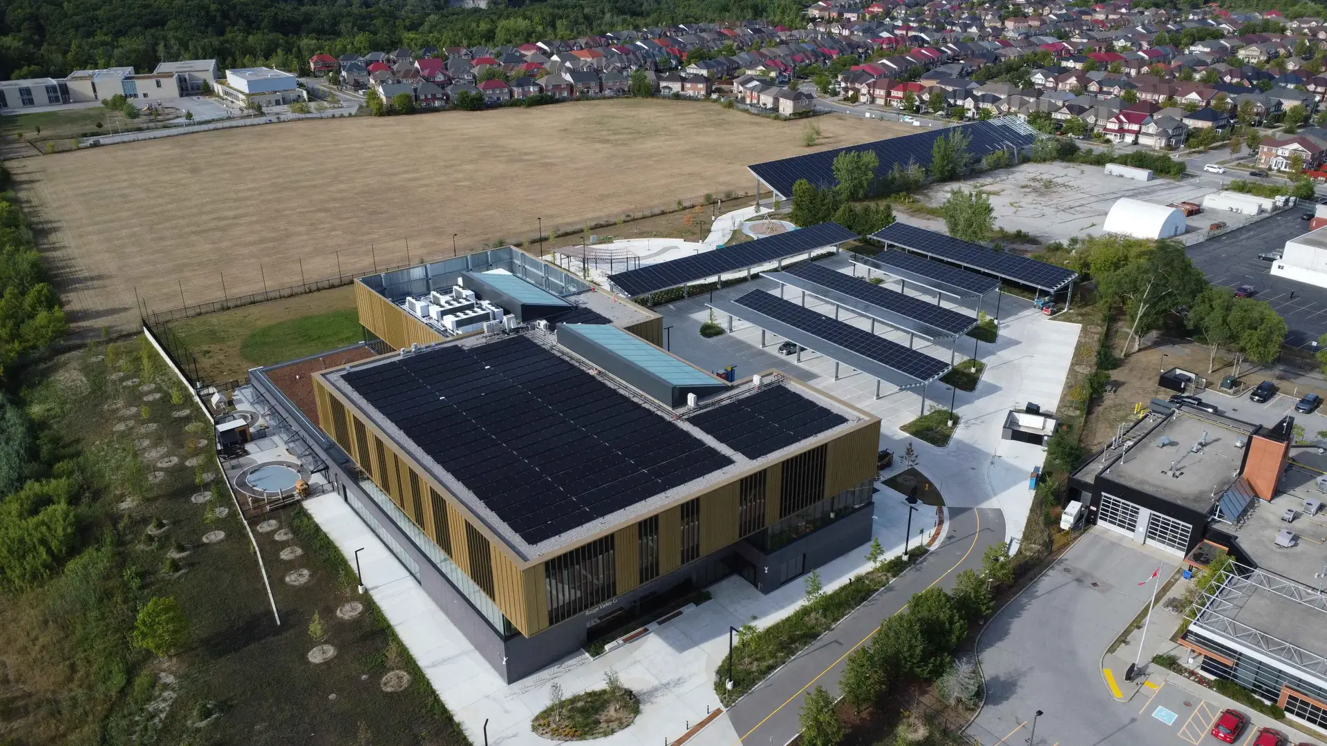 Aerial view of a modern building with solar panels on the roof, adjacent parking, and surrounding fields and houses in the distance.