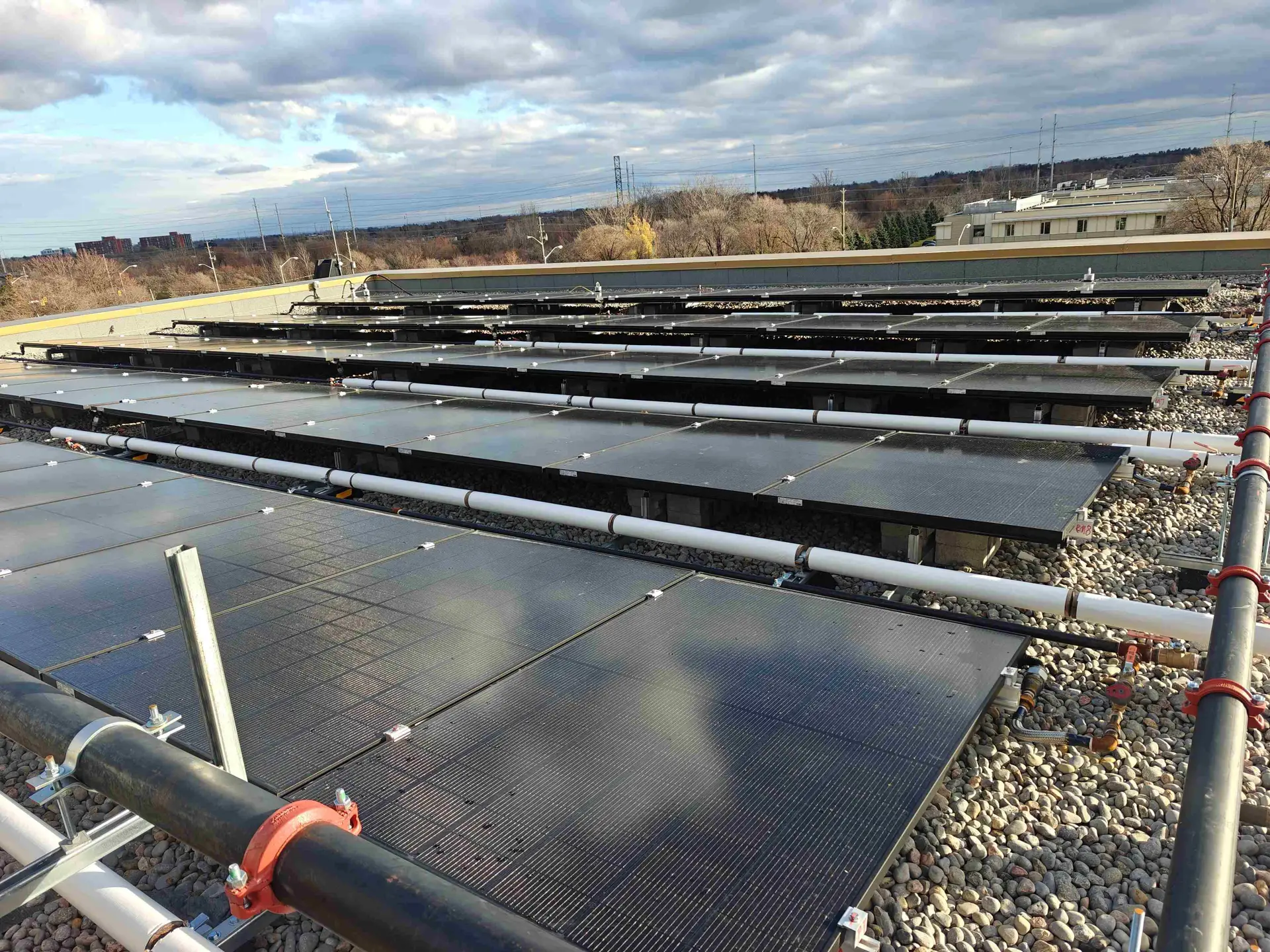 Rooftop solar panels arranged in rows on a gravel roof, with piping and railing in the foreground and a cloudy sky above.