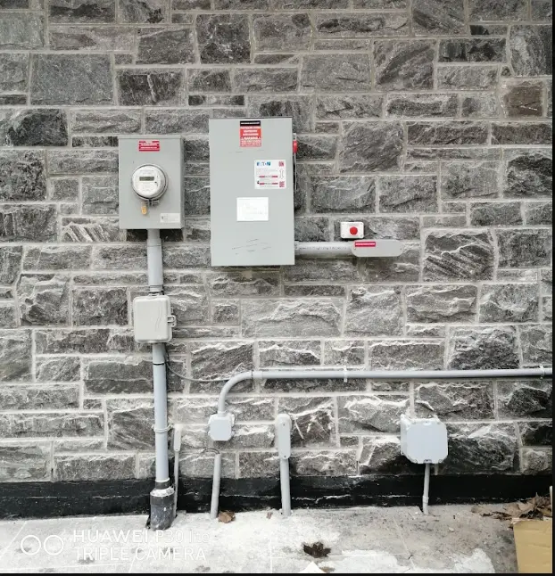 Industrial electrical meters and conduit on a stone wall, with gray control boxes and red safety labels.