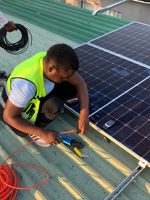 A technician in a bright safety vest works on solar panels on a roof, using tools and cables.
